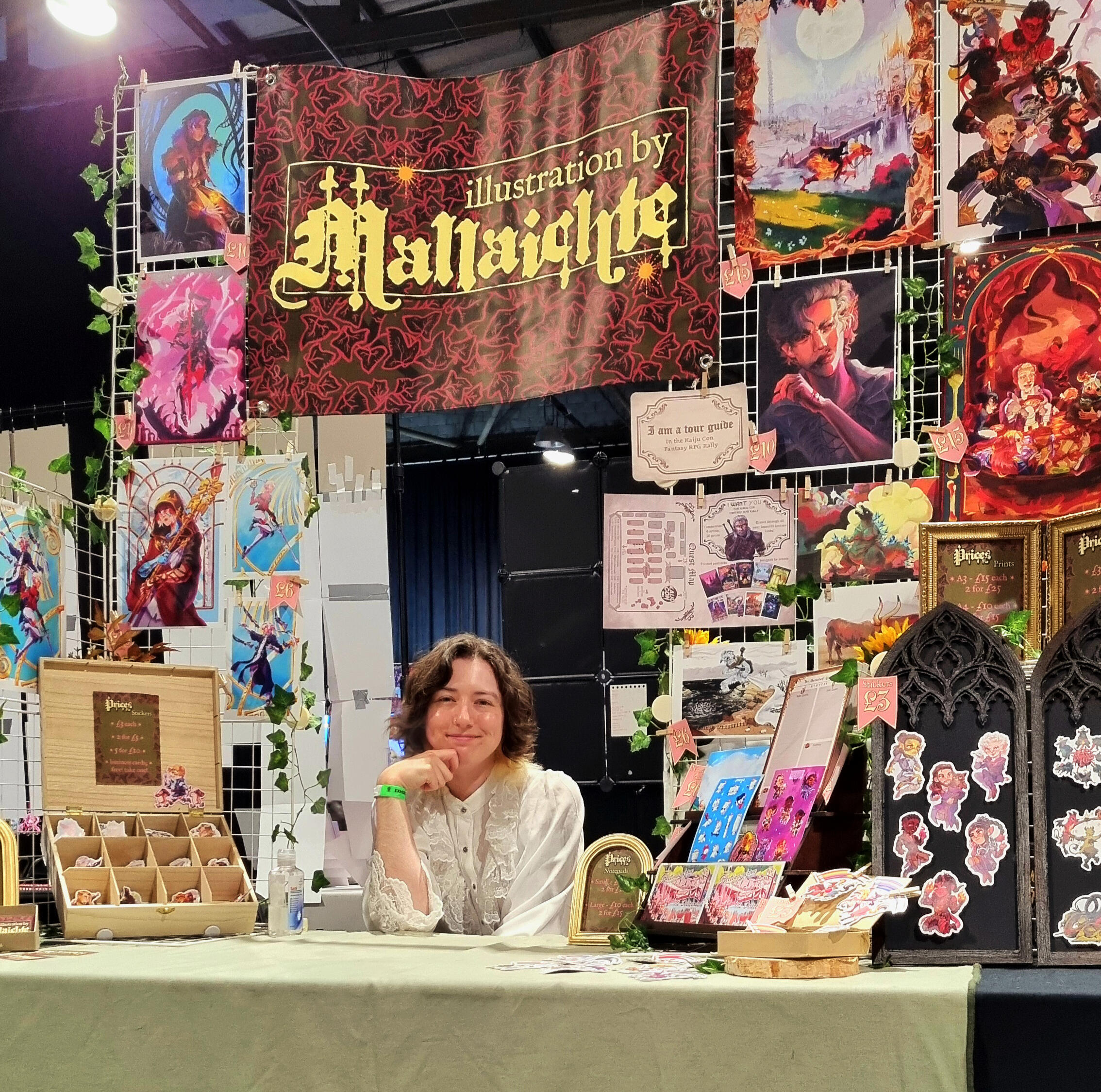 Mal sits behind his artist alley table at KaijuCon 2025. A large sign reading "Illustration by Mallaichte" hangs above a variety of illustrated prints, stickers, and notepads.